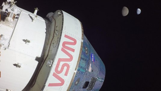 Eine Raumkapsel im All, mit dem Schriftzug NASA an der Außenwand; im Hintergrund ist der Mond und noch weiter entfernt die Erde