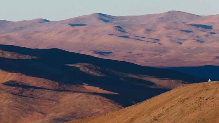 Das Teleskop VISTA in der Atacamawüste von Chile, Bildquelle: ESO/B. Tafreshi (twanight.org); Lizenz: gemäß den Bedingungen der Quelle Vor dem Hintergrund einer bergigen Wüstenlandschaft befindet sich auf einem kleineren Gipfel ein weißes Teleskop-Gebäude.